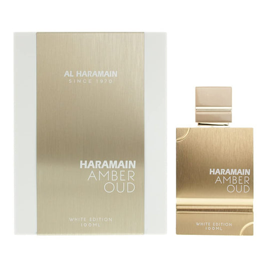 A 100ml bottle and box of Al Haramain Amber Oud White Edition Eau de Parfum showcases a gold-toned design with the brand and product name prominently. The sleek, rectangular bottle is paired with a matching gold cap.