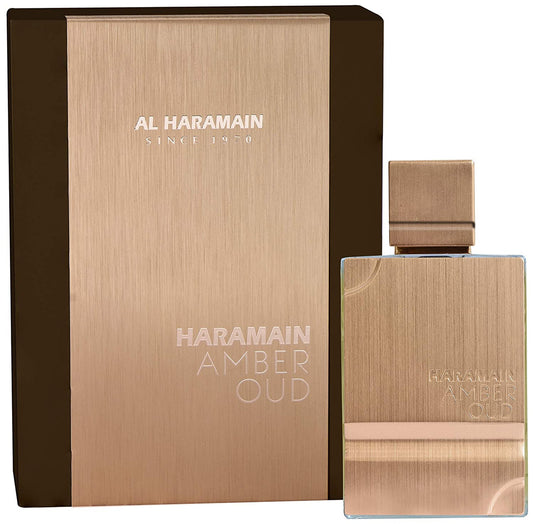 A rectangular, sleek metallic perfume bottle labeled AL HARAMAIN AMBER OUD 2 OZ EDP SP sits beside its matching box, displaying the brand name AL HARAMAIN with Since 1970 above. The overall color is a warm bronze.